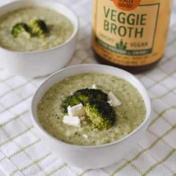 A bowl of creamy broccoli soup topped with cookied broccoli pieces and feta, next to a jar of Zoup Veggie Broth on a white tablecloth.