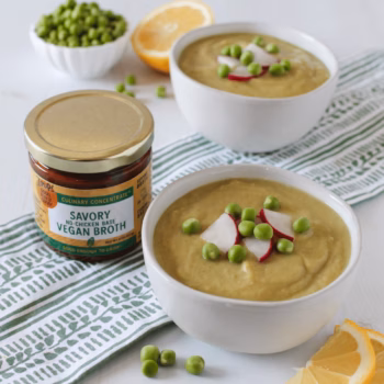 Two white bowls of creamy spring pea soup, garnished with sliced radishes and green peas with a jar of Zoup savory no-chicken base Vegan Broth beside them.