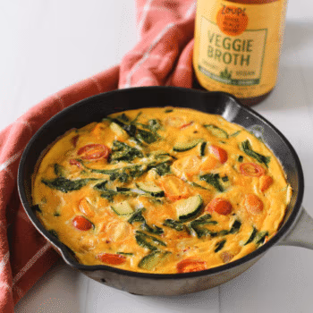 Cast iron skillet with a colorful vegetable frittata made with zucchini, cherry tomatoes, spinach, and red onion, served next to a jar of Zoup Veggie Broth.