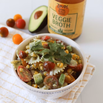 Colorful pasta salad in a white bowl with avocado, cherry tomatoes, corn, and cilantro, set in front of a Zoup Veggie Broth jar and fresh produce.