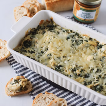 Baked spinach and artichoke dip in a white casserole dish, topped with melted cheese and served with sliced baguette. A jar of Zoup Savory Vegan Concentrate is visible in the background.