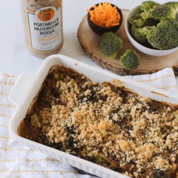 Casserole dish filled with broccoli mushroom bake topped with golden breadcrumbs, made with Zoup Portabella Mushroom Bisque. Ingredients including shredded cheese and fresh broccoli are shown beside the dish.