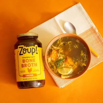 Overhead shot of a jar of Zoup Chicken Bone Broth beside a bowl of lemon orzo chicken soup, with carrots, lemon slices, dill, and orzo, on an orange background.