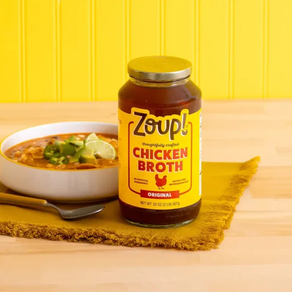 A jar of Zoup Chicken Broth beside a bowl of chicken tortilla soup topped with avocado, jalapeño slices, and lime, on a fringed yellow napkin with a yellow beadboard wall in the background.