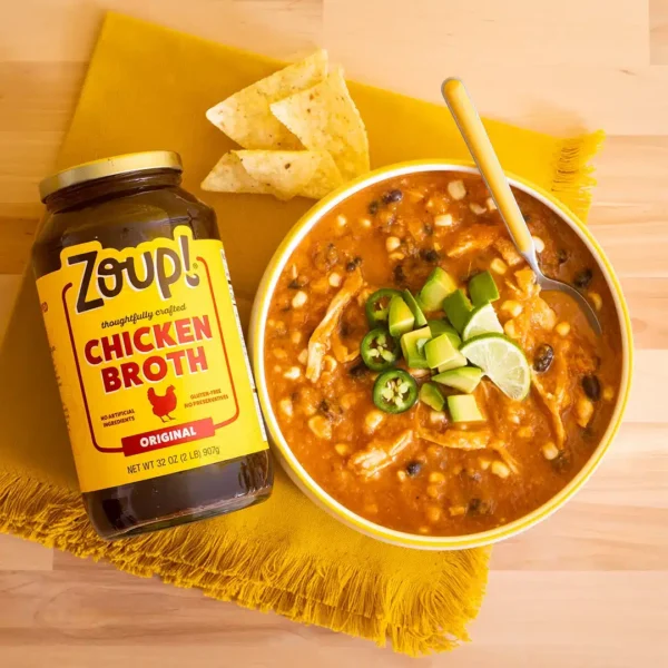 Overhead shot of a jar of Zoup Chicken Broth next to a bowl of chicken tortilla soup topped with avocado, jalapeño slices, lime, and tortilla chips, on a yellow napkin.