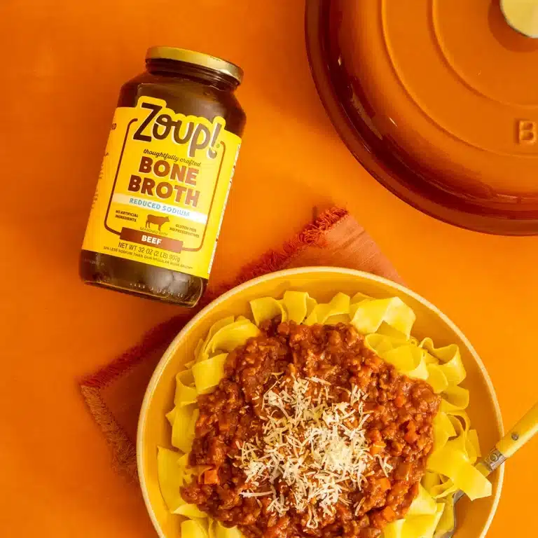 Overhead shot of a jar of Zoup Reduced Sodium Beef Bone Broth beside a bowl of pappardelle topped with beef Bolognese sauce and shredded parmesan, with a Dutch oven in the corner.
