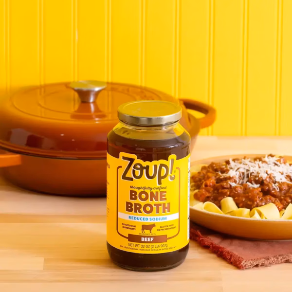 A jar of Zoup Reduced Sodium Beef Bone Broth on a wooden countertop with a plate of pasta topped with beef bolognese sauce and parmesan cheese, and a brown Dutch oven against a yellow background.