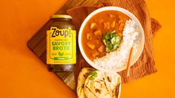Overhead shot of a jar of Zoup Vegan Savory Broth next to a bowl of tofu Tikka Masala with white rice and cilantro, served with naan on a dark wooden board.